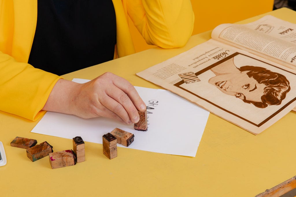 Graafikko Kati leimaa kirjaimia paperille retrolehden äärellä, esimerkkinä käsintehty graafinen suunnittelu.